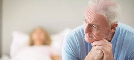 Mann sitzt grübeln am Bettrand, im Hintergrund liegt eine Frau im Bett